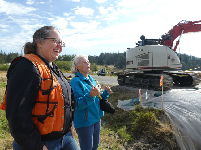 A Female conservation district employee is in the foreground smiling and laughing with an elderly woman with large machinery and work happening in an outdoor setting behind them.